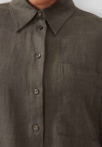 Dark olive green linen shirt with a pointed collar, front buttons, and a single chest pocket. Smooth texture with a natural finish.