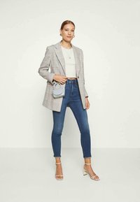 Mulher vestindo um blazer xadrez, top cropped branco, jeans skinny azuis, saltos brancos e segurando uma pequena mala azul clara, de pé contra um fundo simples.