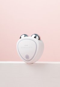 Stylpro Gesichtsmassager aus weißem Kunststoff in Herzform, mit metallischen Kugeln an der Spitze und einem lila Einschaltknopf. Minimalistisches Design.