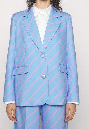 Frau trägt einen hellblauen Anzug mit pinkfarbenen diagonalen Streifen und floralen Knöpfen über einem weißen Hemd mit Kragen, gezeigt vom Hals bis zu den Oberschenkeln.