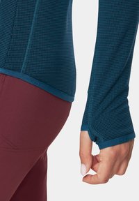 Długi rękaw koszuli w ciemnym teal z fakturowanym wzorem, z otworami na kciuki przy mankietach i niebieskim wykończeniem wzdłuż dołu. Zestawione z bordowymi spodniami.