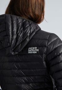 Svart puffjacka med quiltat mönster och huva. Har en vit logotyp "JACK1T SATELITE DES1GN" på vänster axel. Slät textur.