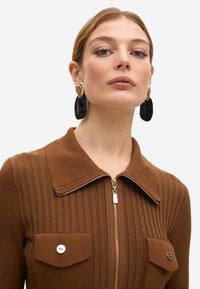 Top marrón con canalé, con cierre frontal, mangas largas y gran collar. Cuenta con dos bolsillos delanteros con botones y detalles en tono dorado.