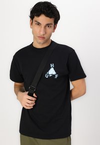 Black cotton t-shirt featuring a white and blue triangular logo with "HUF" lettering. Model holds a black shoulder strap bag.