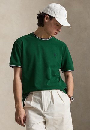 Jeune homme portant une chemise verte à manches courtes avec une bordure bleu marine, pantalon blanc, casquette blanche et montre-bracelet, regardant vers le bas à sa gauche.