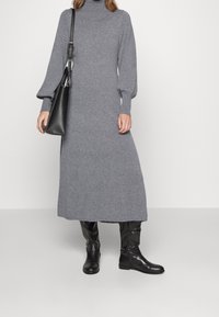 Šedé pletené šaty s vysokým límcem a dlouhými nabíranými rukávy, doplněné černými kozačkami po kolena a černou kabelkou přes rameno. Hladká textura.
