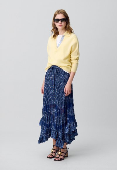 Pullover in maglina giallo chiaro con scollatura a V, sovrapposto a una gonna blu a strati con motivo. Occhiali da sole neri e décolleté con cinturini e dettagli dorati.