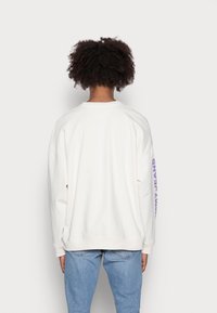 Felpa oversized color crema con orlo e polsini a costine. Presenta un testo viola sulla manica destra. Indossata con jeans azzurri chiari.