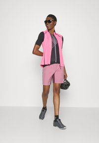 Roze mouwloze vest over een zwart t-shirt met korte mouwen, gecombineerd met roze shorts en grijze sportschoenen. Zwarte zonnebril en helm inbegrepen.