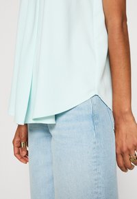 Haut sans manches bleu clair à la coupe fluide, doté de plis latéraux et d'un ourlet asymétrique. Porté avec un jean en denim bleu clair.