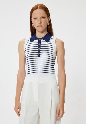 Kobieta nosząca biały, bezrękawnikowy top dzianinowy z granatowymi paskami i kołnierzykiem, w połączeniu z białymi spodniami z wysokim stanem, zwrócona przodem.