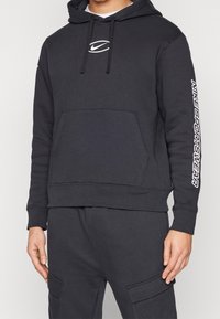 Svart huva sweatshirt i bomullsblandning, med en framficka i kängurustil, ribbade muddärmar och "Nike Sportswear" tryckt på ärmen.