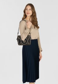 Zwarte handtas met ingewikkelde bloemmotieven van kralenwerk, zilveren studs en een afneembare riem, gecombineerd met een beige blouse en een geplooide marineblauwe rok.