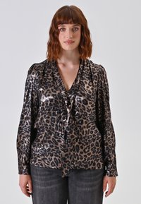 Donna con capelli ondulati di lunghezza spalla indossa una blusa lucida con stampa leopardo, con un collo a V legato, e jeans scuri, in piedi di fronte a uno sfondo semplice.