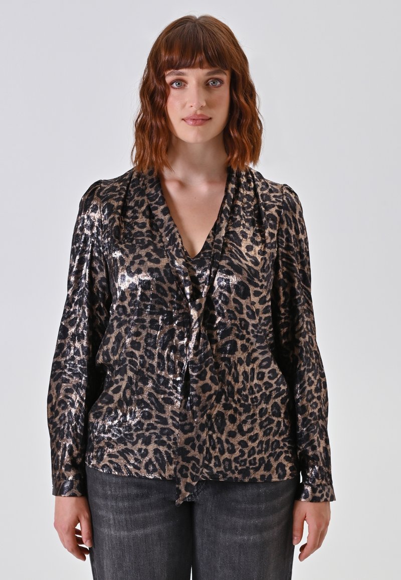 Donna con capelli ondulati di lunghezza spalla indossa una blusa lucida con stampa leopardo, con un collo a V legato, e jeans scuri, in piedi di fronte a uno sfondo semplice.