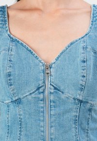 Denim corset top features a deep V neckline and a silver zipper at the center. The fabric is light blue with a smooth texture and structured seams.