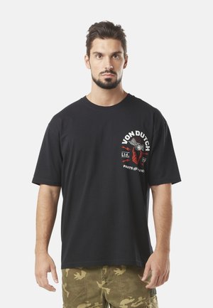 Homme avec une barbe portant un t-shirt graphique noir Von Dutch et un short cargo camouflage, se tenant devant un fond blanc.