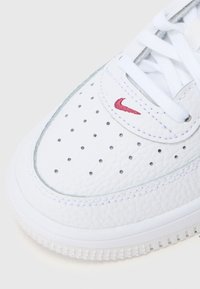 Balti ādas sneakeri ar nelieliem ventilācijas caurumiem uz purngalu, baltiem korsetes saistēm un sarkanu izšūtu Nike swoosh logotipu uz priekšējā paneļa.