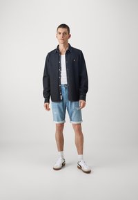 Chemise en denim sombre, débardeur blanc, shorts déchirés bleu clair, baskets blanches et un collier en perles. Tenue simple et décontractée.