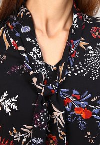 Blusa nera con un motivo floreale nei toni del rosso, blu e crema, caratterizzata da un collo legato e un tessuto leggero e morbido con una consistenza liscia.
