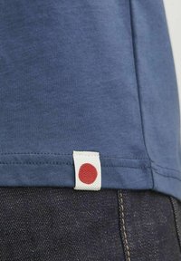 Un tricou din bumbac albastru cu un etichetă logo sub formă de punct roșu aproape de tiv, contrastând cu blugi din denim închisi la culoare care au detalii de cusătură vizibile.