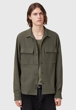 Olive green button-up shirt, long sleeves, two front pockets, flat collar, textured fabric, worn over a matching shirt, part of a casual outfit.