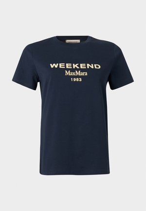 Femme debout, une main dans la poche, portant un t-shirt noir "Weekend Max Mara", un jean large bleu et des chaussures noires à bout pointu.