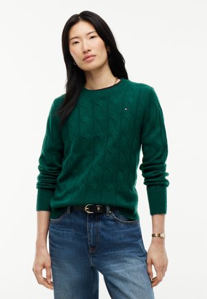 Donna con lunghi capelli neri che indossa un maglione verde a trecce e jeans blu su uno sfondo bianco uniforme.