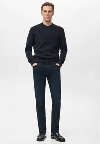 Suéter de punto azul marino con cuello redondo, combinado con pantalones oscuros de corte ajustado y zapatos de cuero negros. El conjunto general es minimalista.