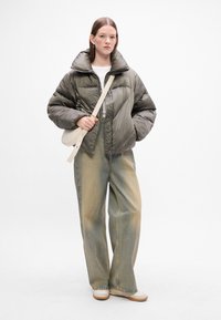 Olivengrønn pufferjakke med høy krage, glidelås foran og sidelommer, kombinert med lyseblå vide jeans og beige sko.