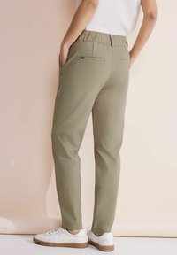 Pantaloni kaki con cintura elastica, due tasche laterali e due tasche posteriori a soffietto, abbinati a sneakers bianche, texture liscia.