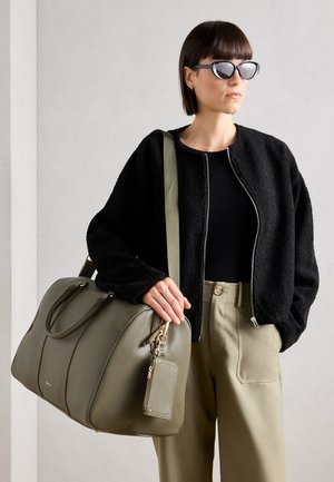 Borsa in pelle verde con forma arrotondata, doppio manico e tracolla removibile. Una giacca nera strutturata e pantaloni beige completano il look.