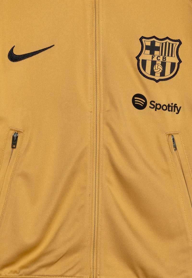 Chaqueta amarilla de Nike con el logo negro de FCB y el emblema de Spotify. Cuenta con bolsillos con cremallera y cierre completo. Está hecha de un tejido suave.