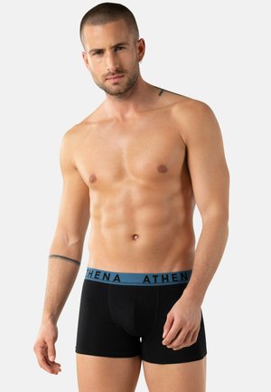 Homme athlétique torse nu portant un boxer noir avec une ceinture bleue marquée "ATHENA", debout devant un fond clair uni.