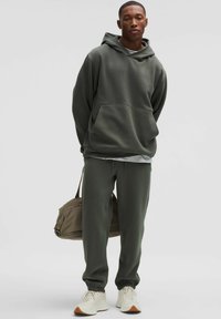 Olívazöld kapucnis pulóver, nagy elülső zsebbel, amelyhez passzoló melegítőnadrág tartozik. Lazább szabású és puha anyagból készült, fehér sportcipővel párosítva.