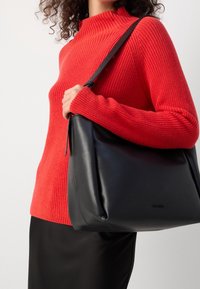 Zwarte leren schoudertas met een gladde textuur, voorzien van een brede opening en een enkele schouderband, gecombineerd met een rood geribd sweater.