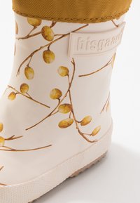Gumowa kalosza z błyszczącym kremowym wykończeniem, ozdobiona wzorem gałązki żółtej jagody. Teksturowana brązowa podeszwa i beżowy materiałowy mankiet.