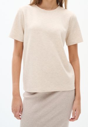 Frau trägt ein beiges, kurzärmeliges Rundhalsshirt und einen gerippten, beigen Rock, steht mit locker an den Seiten hängenden Armen vor weißem Hintergrund.