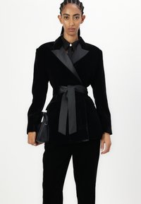 Black velvet blazer with satin accents, tie belt, notched collar, long sleeves, and paired with black trousers and small handbag.