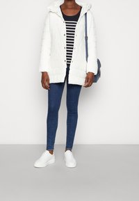 Chaqueta de plumas blanca con capucha, textura acanalada y botones frontales, sobre una camiseta de rayas azul y blanca, combinada con jeans azules y zapatillas blancas.