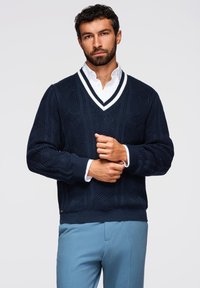 Granatowy sweter z dzianiny z dekoltem w serek i białymi wykończeniami, z fakturowanymi wzorami. Noszony na białej koszuli z kołnierzykiem i zestawiony z jasnoniebieskimi spodniami.