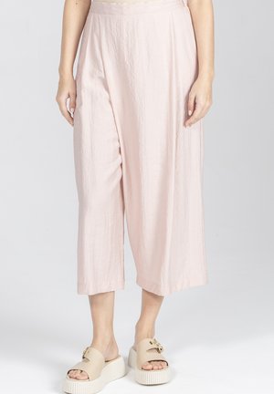 Kobieta w jasnoróżowych, szerokich, krótkich spodniach i beżowych sandałach na platformie z złotą klamrą, stojąca na jednolitym jasnym tle.