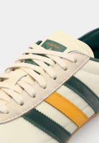 Adidași cu un design crem, având dungi verzi și portocalii, accente din piele întoarsă și șireturi bej. Talpa din cauciuc texturat este vizibilă.