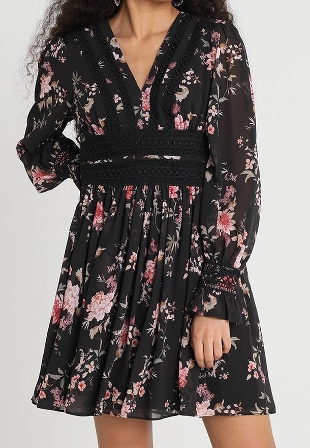 Femme portant une robe noire fleurie avec des manches longues transparentes, une bordure en dentelle sur les manches et la taille, et un décolleté en V.