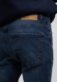 Jeans de mezclilla azul oscuro con una textura suave, que cuentan con dos bolsillos traseros y una etiqueta de cuero marrón en la cintura con texto en relieve.