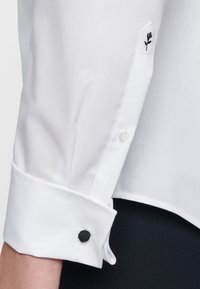 Vit skjorta med knappar som har en svart broderad blommönster på ärmen och en kontrasterande svart knapp på manschetten.