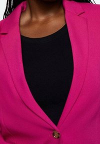 Personne portant un blazer rose vif par-dessus un haut noir, montrant le contour du cou et le haut de la poitrine avec un seul bouton fermé sur le blazer.