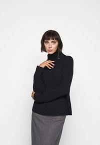 Pull en mailles côtelées noir à col roulé, à manches longues, avec des fentes latérales, associé à une jupe à motif pied-de-poule.