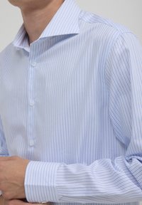Uomo che indossa una camicia a maniche lunghe con righe azzurre e bianche, mentre sistema il polsino con la mano sinistra visibile.