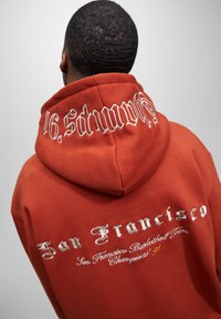 Sudadera naranja con capucha ajustable, con texto bordado en blanco y plata en la parte posterior, incluyendo "San Francisco" y detalles deportivos.